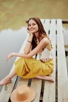 une hippie femme est assis avec sa chapeau sur une pont par une Lac avec sa mains en haut dans le air sur une la nature voyage et souriant Heureusement dans éco-vêtements. détendu mode de vie photo