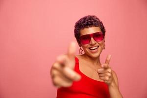 mode portrait de une femme avec une court la Coupe de cheveux dans coloré des lunettes de soleil avec inhabituel accessoires avec des boucles d'oreilles sourit sur une rose brillant Contexte spectacles sa mains à le caméra photo