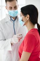 une patient étant examiné par une médecin portant une médical masque, protecteur gants et une stéthoscope autour sa cou photo