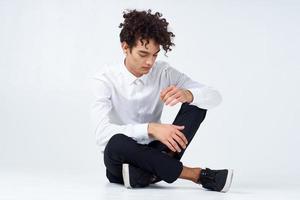 une gars avec frisé cheveux dans une blanc chemise, baskets et pantalon est assis sur le sol dans une brillant pièce photo