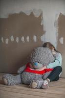 une triste neuf ans fille, elle est assis dans une pièce dans le grenier sans pour autant réparations sur le sol, étreint un vieux gros nounours ours, enfants angoisses et chagrins photo