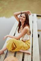 une hippie femme est assis avec sa chapeau sur une pont par une Lac avec sa mains en haut dans le air sur une la nature voyage et souriant Heureusement dans éco-vêtements. détendu mode de vie photo