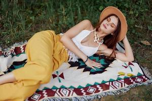 une femme dans respectueux de la nature Vêtements dans une hippie Regardez est mensonge sur une coloré plaid souriant et à la recherche à un l'automne le coucher du soleil dans la nature dans le parc. mode de vie sur une périple de harmonie avec le monde photo