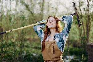 une femme souriant magnifiquement et à la recherche à le caméra, une agriculteur dans travail vêtements et un tablier travail en plein air dans la nature et en portant une râteau à recueillir herbe et fourrage pour le animaux dans le jardin photo