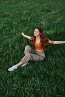 une femme profiter le en plein air séance dans le parc sur le vert herbe dans décontractée Vêtements avec longue écoulement cheveux, allumé par le brillant été Soleil sans pour autant les moustiques photo