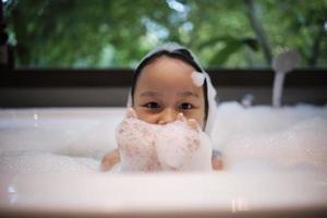 portrait de un asiatique peu mignonne fille avec bonheur et amusement en jouant avec corps savon mousse, shampooing éclaboussure, et l'eau bulles dans une blanc baignoire à une Hôtel salle de bains avec vert arbre fenêtre Contexte. photo