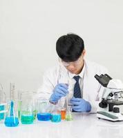 asiatique Masculin étudiant scientifique portant une médecins robe dans le laboratoire à la recherche main à chimiste. causé par mélange réactifs dans scientifique recherche laboratoires avec tester tubes et microscope sur le table photo