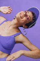 portrait de une sportif mode femme posant souriant avec les dents à le caméra avec élégant des boucles d'oreilles dans sa oreilles dans une violet yoga survêtement et une transparent casquette sur une violet monochrome Contexte photo