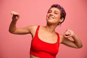 mode portrait de une femme avec une court la Coupe de cheveux de violet Couleur et une sourire avec les dents dans une rouge Haut sur une rose Contexte dansant Heureusement photo