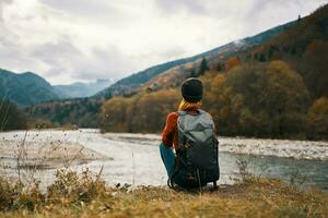 femme avec sac à dos sur le rivière banque admire le Montagne paysage dans le Contexte photo