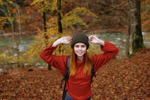 joyeux femme dans une chapeau chandail avec une sac à dos sur sa retour gestes avec sa mains dans une parc dans la nature photo