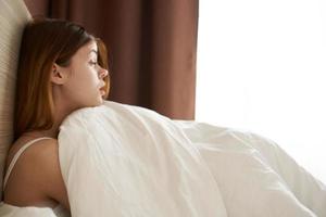 une jolie femme dans une nuit costume est mensonge dans lit en dessous de une couverture près le fenêtre sur une fin de semaine vacances photo