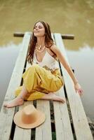 une hippie femme est assis avec sa chapeau sur une pont par une Lac avec sa mains en haut dans le air sur une la nature voyage et souriant Heureusement dans éco-vêtements. détendu mode de vie photo