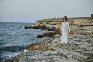 jolie femme dans blanc robe des stands sur le des pierres plage la nature liberté inchangé photo