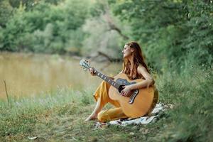 hippie femme en jouant guitare souriant et en chantant Chansons dans la nature séance sur une plaid par le Lac dans le soir dans le des rayons de le réglage Soleil. une mode de vie dans harmonie avec le corps et la nature photo