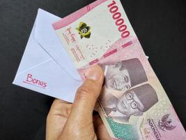 homme en portant une blanc enveloppe écrit de prime et Nouveau indonésien billets de banque, d'habitude Tunjangan hari raya ou appelé thr sont donné à des employés devant de aïd. isolé sur noir Contexte et Haut vue photo