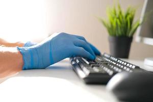 Masculin mains dans bleu nitrile gants utilisation le clavier sur le ordinateur, faire le travail. Bureau travail dans le covid-19 pandémie. photo