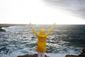 Jeune femme vêtue d'un imperméable jaune debout avec les bras tendus tout en profitant du magnifique paysage de mer un jour de pluie sur une plage rocheuse par temps nuageux au printemps photo