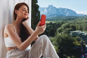 attrayant Jeune femme utilisation le téléphone regards à le écran dans une confortable Hôtel un ouvert vue de le Montagne vue photo