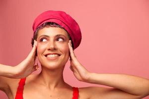 Jeune athlétique femme avec une court la Coupe de cheveux et violet cheveux dans une rouge Haut et une rose chapeau avec un athlétique figure sourit et grimaces à la recherche à le caméra sur une rose Contexte photo