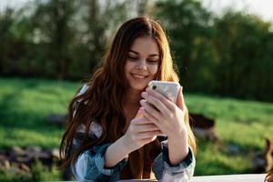 une magnifique femme relaxant et travail sur sa téléphone séance dans la nature dans le parc parmi le des arbres souriant et en portant sa téléphone intelligent dans sa main allumé par le brillant le coucher du soleil lumière photo