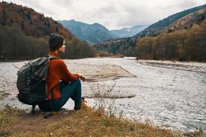 femme avec une sac à dos dans une veste et une chapeau sur le rivière banque dans le montagnes côté vue photo