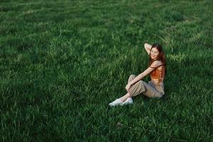 une femme profiter le en plein air séance dans le parc sur le vert herbe dans sa décontractée vêtements allumé par le brillant été Soleil sans pour autant les moustiques photo