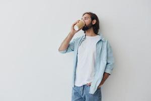 free-lance millénaire homme avec une barbe en buvant café de une recyclé tasse dans élégant branché vêtements blanc T-shirt bleu jeans et chemise sur une blanc Contexte photo