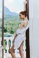 portrait de une magnifique femme dans une blanc chemise admire le vert la nature sur le balcon inchangé photo