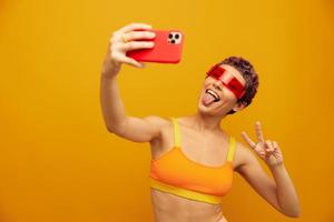 femme sourit et spectacles sa langue dans inhabituel des lunettes millénaire prend selfies sur sa téléphone dans tenue de sport contre un Orange studio toile de fond, gratuit espace photo