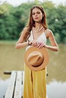 une Jeune femme souriant dans un image de une hippie et éco-robe dansant dans la nature par le Lac portant une chapeau et Jaune un pantalon dans le été le coucher du soleil photo