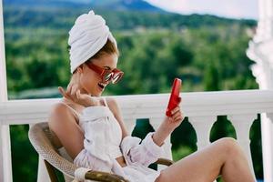 attrayant Jeune femme dans des lunettes de soleil est assis sur le balcon et les usages le téléphone paresseux Matin photo