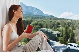 portrait de magnifique femme utilisation le téléphone regards à le écran dans une confortable Hôtel un ouvert vue de le inchangé photo