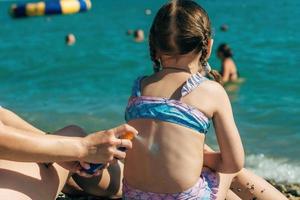 maman soigneusement frottis sa enfant peau avec une protecteur crème sur le plage. peau se soucier et Soleil protection. crème solaire vaporisateur pour les enfants. photo