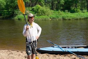 une homme avec une kayak pagayer pour rafting des stands sur le rivière banque. sport l'eau randonnée, une été aventure. respectueux de la nature et extrême tourisme, actif et en bonne santé mode de vie photo