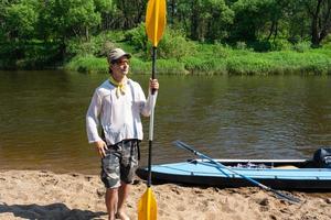 une homme avec une kayak pagayer pour rafting des stands sur le rivière banque. sport l'eau randonnée, une été aventure. respectueux de la nature et extrême tourisme, actif et en bonne santé mode de vie photo