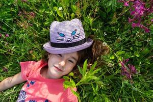 portrait de une enfant dans une chapeau avec le sien visage couvert dans été mensonge dans le herbe et fleurs sauvages. chapeau avec yeux et oreilles comme une chat, été temps, liberté photo