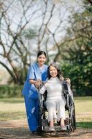 asiatique prudent soignant ou infirmière et le content patient dans une fauteuil roulant sont en marchant dans le jardin. à Aidez-moi et encourager et du repos votre esprit avec vert la nature. utilisation une stéthoscope à Ecoutez à votre photo