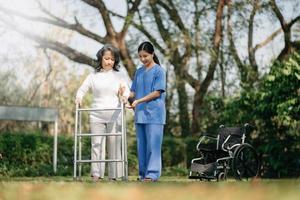 asiatique prudent soignant ou infirmière et le content patient dans une fauteuil roulant sont en marchant dans le jardin à Aidez-moi et encourager et du repos votre esprit avec vert la nature. Aidez-moi soutien toi même à apprendre à marcher. photo