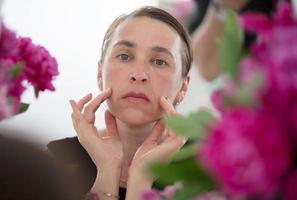 une milieu vieilli femme examine les rides contre une Contexte de fleurs. se soucier pour vieillissement peau. photo