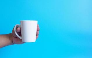 main d'homme tenant une tasse de café blanche, remplie de café noir chaud americano, sans sucre ni lait, prêt à boire, rafraîchissant. arôme éveillé frais au travail placé sur un fond bleu isolé photo