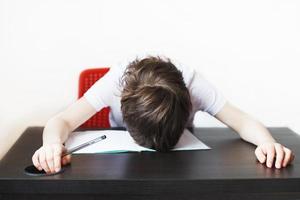 le garçon est fatigué de Faire devoirs. enfant est assis et Est-ce que devoirs sur une blanc Contexte photo