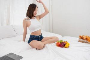 portrait de blanc asiatique femme avec une tatouage séance sur le lit relaxant et en jouant portable sur fin de semaine là a été pain et fruit et rouge pommes sur une blanc lit à manger. vacances concept photo