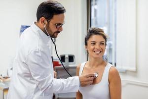 Jeune médecin Ecoutez à femelle patient cœur poitrine avec stéthoscope à clinique réunion. homme gp vérification examiner femme client avec phonendoscope. soins de santé concept. photo