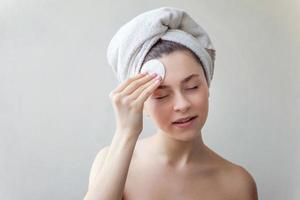 femme souriante dans une serviette sur la tête avec une peau douce et saine enlevant le maquillage avec un tampon de coton isolé sur fond blanc photo