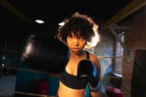 femmes soi la défense pouvoir. africain américain femme combattant perforation avec boxe gants à caméra. en bonne santé fort fille perforation formation poinçons à la recherche concentré droit. en forme corps entraînement. photo