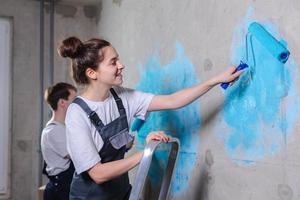 maison décoration DIY rénovation concept. content couple dans vide pièce de Nouveau Accueil La peinture mur ensemble. mari et épouse en portant peindre rouleau La peinture mur avec bleu Couleur peindre dans Nouveau maison. photo