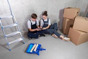 maison DIY rénovation concept. Jeune content couple séance sur sol à la recherche Accueil réparation plan dans tablette à l'intérieur. femme montrant à homme plan intérieur conception en ligne. couple dans Nouveau Accueil pendant réparation travaux. photo