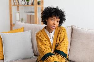 belle fille afro-américaine avec une coiffure afro souriante assise sur un canapé à la maison à l'intérieur. jeune femme africaine aux cheveux bouclés en riant. liberté bonheur insouciant concept de gens heureux. photo