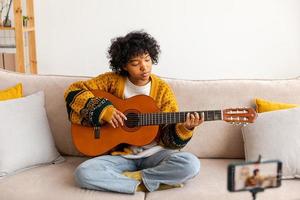 blogueur guitariste. content africain américain fille blogueur en jouant guitare en chantant chanson enregistrement vlog. social médias influenceur femme diffusion enregistrement à Accueil studio. la musique contenu créateur diffuser. photo